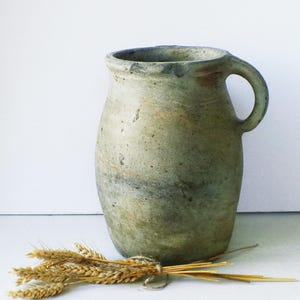 May include: A rustic, pale green ceramic jug with a handle. The jug has a textured surface and a wide opening. A small bundle of wheat stalks rests in front of the jug.