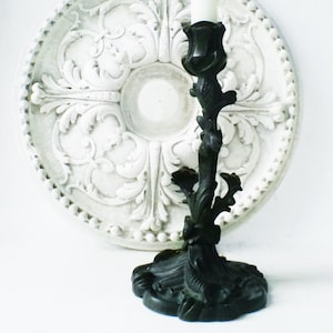 May include: A black, ornate candlestick holder with a floral design. The candlestick is made of metal and has a detailed, textured surface. A white candle sits in the holder. The background features a white, decorative wall medallion.