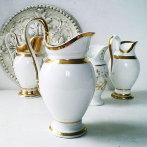 May include: A collection of white porcelain pitchers with gold trim. The pitchers vary in size and design, with curved handles and decorative gold accents around the rims and bases. A decorative plate is in the background.
