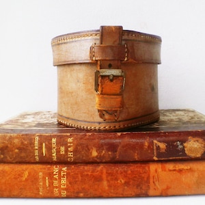 Puede incluir: Una sombrerera cilíndrica de cuero vintage con cierre de hebilla, colocada sobre dos libros antiguos. Los libros tienen cubiertas envejecidas y texturizadas en tonos marrones y naranjas, con letras doradas visibles en los lomos. La sombrerera es de color marrón claro.