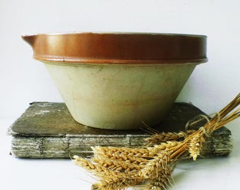 May include: A vintage stoneware mixing bowl with a brown rim and a light green body. The bowl sits on a weathered book, with a bundle of wheat tied with twine. The bowl has a spout for pouring.