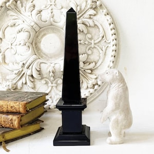 Könnte beinhalten: Eine schwarze Obelisk-Skulptur mit quadratischem Sockel, die auf einer weißen Oberfläche steht. Der Obelisk steht neben einer kleinen, weißen Eisbärfigur. Antike Bücher mit vergilbten Seiten befinden sich im Hintergrund.