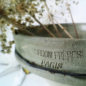 Peut inclure: Gros plan sur un bol en céramique grise texturée avec le texte "OURDON FRERES PARIS" gravé sur le côté. Le bol contient des tiges de plantes séchées de couleur beige. Le bol a un bord foncé.