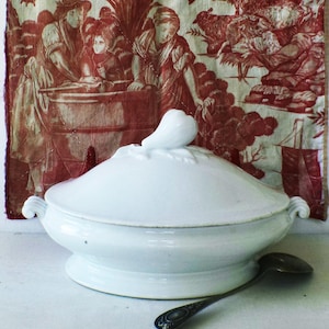 May include: White ceramic soup tureen with a lid and decorative handle. The tureen has side handles and sits on a pedestal base. A silver spoon rests beside it. The background features a red and white toile fabric.