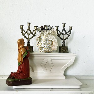 Puede incluir: Una exhibición vintage con una figura arrodillada en rojo y marrón, flanqueada por dos candelabros de latón ornamentados. Un jarrón floral blanco y dorado con adornos de encaje y elementos decorativos se encuentra en un estante blanco.