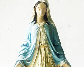 May include: A religious statue of a figure with dark hair, wearing a white head covering and a light blue cloak over a cream-colored gown. The cloak features gold-colored trim along the edges, and the figure has hands extended outward.