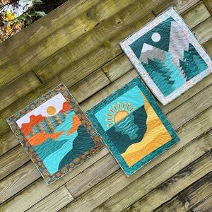 May include: Three colorful quilted wall hangings with nature scenes. The first features a mountain range with a full moon, the second has a sun setting over a lake, and the third shows a sunset over a mountain range.