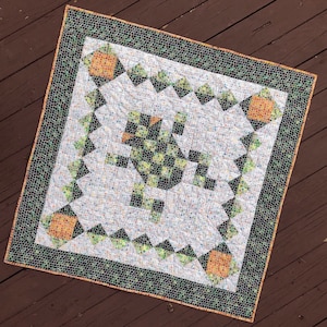 Puede incluir: Una colcha blanca con un patrón de cuadros verdes y negros en el centro. La colcha está rodeada por un borde de mosaicos verdes y marrones. La colcha está sobre una superficie de madera.