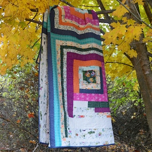 May include: A colorful quilt with a log cabin pattern hangs from a tree branch. The quilt is made of various fabrics in shades of blue, green, purple, pink, and orange. The background is a blurred image of fall foliage.