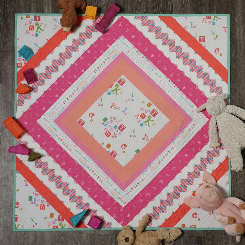 May include: A pink and white quilt with a diamond-shaped center featuring a white background with colorful letters and illustrations. The quilt is surrounded by a pink and white border with a scalloped edge. The quilt is on a light brown surface with toys scattered around it.