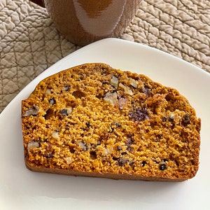 May include: A slice of golden-brown bread with visible nuts and berries on a white plate, next to a brown mug filled with dark coffee. The bread has a textured surface, and the coffee mug has a handle.