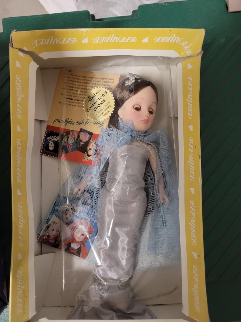 Puede incluir: Una mu&ntilde;eca Effanbee vintage en su caja original. La mu&ntilde;eca viste un vestido plateado, una capa de velo azul y un collar de perlas. La caja es amarilla con el logo de Effanbee y las palabras "Collector's Choice".