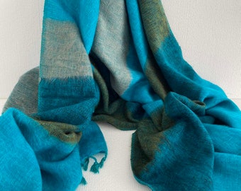 May include: A woven scarf featuring a large-scale plaid pattern in vibrant shades of turquoise, teal, and muted olive green. The fabric has a visible textured weave and frayed edges at the ends, creating a soft, draped appearance with varied color blocks.