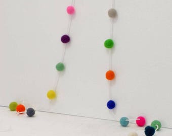 Puede incluir: Una guirnalda colorida de bolas de fieltro enhebradas en un hilo blanco. La guirnalda está colgada en una pared blanca y presenta una variedad de colores, incluyendo rojo, naranja, amarillo, verde, azul, morado, rosa y gris.