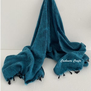 May include: A teal blue pashmina scarf with black fringe and a subtle pattern. The scarf is folded and draped over a white surface. The text "Cashmere Crafts" is visible in the bottom right corner of the image.