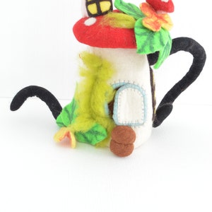 May include: A felt mushroom house with a red roof, white walls, and a green door. The house has a black felt spout and a green felt leaf with a yellow flower.