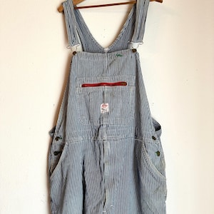 Puede incluir: Overol a rayas azules y blancas con tirantes ajustables y un bolsillo delantero. El overol tiene un bolsillo con cremallera rojo y está colgado de una percha de madera. El diseño general es de estilo workwear clásico.
