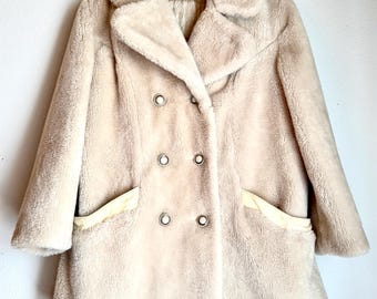 May include: A vintage beige faux fur coat with a double-breasted design. The coat features a wide collar, two front pockets, and six decorative buttons. The coat is hanging on a wooden hanger.