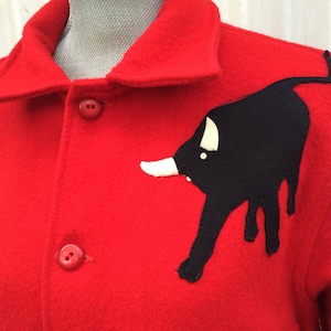 May include: A bright red wool coat with a black bull applique on the front. The bull has white horns and a white spot. The coat has a collar and red buttons.