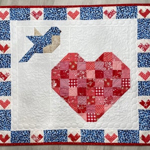 May include: A white quilted fabric wall hanging with a blue and red border. The center features a patchwork heart design with red, white, and blue squares. A blue bird with white accents is in the upper left corner.