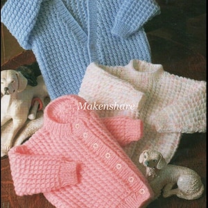 Op de afbeelding: Drie gebreide baby-vestjes in verschillende kleuren: lichtblauw, roze en wit met roze stippen. De vestjes liggen op een houten oppervlak met twee kleine witte keramische hondjes.