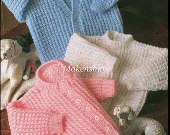 Könnte beinhalten: Drei gestrickte Baby-Cardigans in verschiedenen Farben: hellblau, rosa und weiß mit rosa Sprenkeln. Die Cardigans liegen auf einer Holzoberfläche mit zwei kleinen weißen Keramik-Hunden.