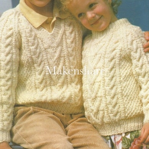 May include: Two children wearing cream colored cable knit sweaters. The sweater on the left has a V-neck and the sweater on the right has a round neck.
