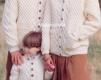 May include: Cream-colored cable-knit cardigan sweaters with brown buttons are worn by a family. The sweaters have pockets and a textured diamond pattern. The family is standing outdoors, with the child in the foreground.
