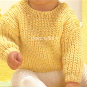 May include: A pale yellow knitted baby sweater with long sleeves and a round neckline. The sweater features three small, light blue buttons on each shoulder. The text "Makenshare" is visible on the sweater.