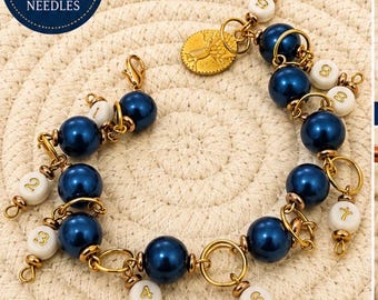 Könnte beinhalten: Ein Set aus Strick-Maschenmarkierern mit dunkelblauen Perlen, weiß nummerierten Perlen von 1 bis 9 und goldfarbenen Metallringen. Ein goldfarbener runder Anhänger mit Prägung ist befestigt. Der Text auf dem Bild lautet: FITS UP TO 8 MM NEEDLES.