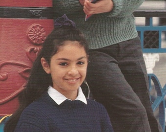 May include: Two children wearing dark blue and green knit sweaters, with white collared shirts underneath. One child is writing on a notepad with a pencil, while the other holds a notepad and pencil. The background includes a red and blue painted structure.