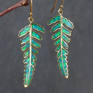 Könnte beinhalten: Goldfarbene Ohrringe in Form von Farnblättern. Die Blätter sind mit einem schimmernden grün-blauen Material gefüllt, das Opal ähnelt. Die Ohrringe hängen an zarten goldenen Haken und werden auf einem kleinen Ast vor grauem Hintergrund präsentiert.
