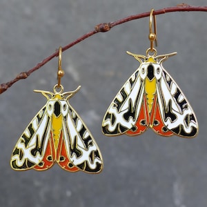 May include: A pair of gold-toned metal earrings shaped like moths. Each moth features enamel detailing in white, black, yellow, and orange. The earrings hang from gold-toned hooks and are suspended from a thin, brown, textured twig.
