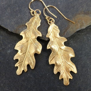 May include: A pair of gold-toned drop earrings shaped like oak leaves. Each earring features a textured surface with a prominent central vein and scalloped edges, suspended from a simple gold-toned wire hook for pierced ears.