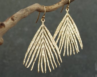 May include: A pair of gold-toned dangle earrings featuring a fan-shaped palm leaf design. Each earring has a textured, hammered surface and is suspended from a simple gold-toned hook wire. The metal pieces are shaped like thin, radiating fronds.