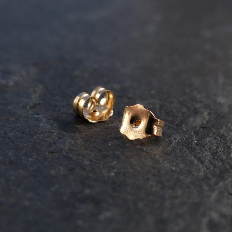 Op de afbeelding: Twee goudkleurige oorbelachterkantjes in de vorm van een bloem. Een van de achterkantjes is zo geplaatst dat de binnenkant zichtbaar is, terwijl de andere schuin staat om de buitenkant te tonen, gestempeld met de tekst 1/20 14K.