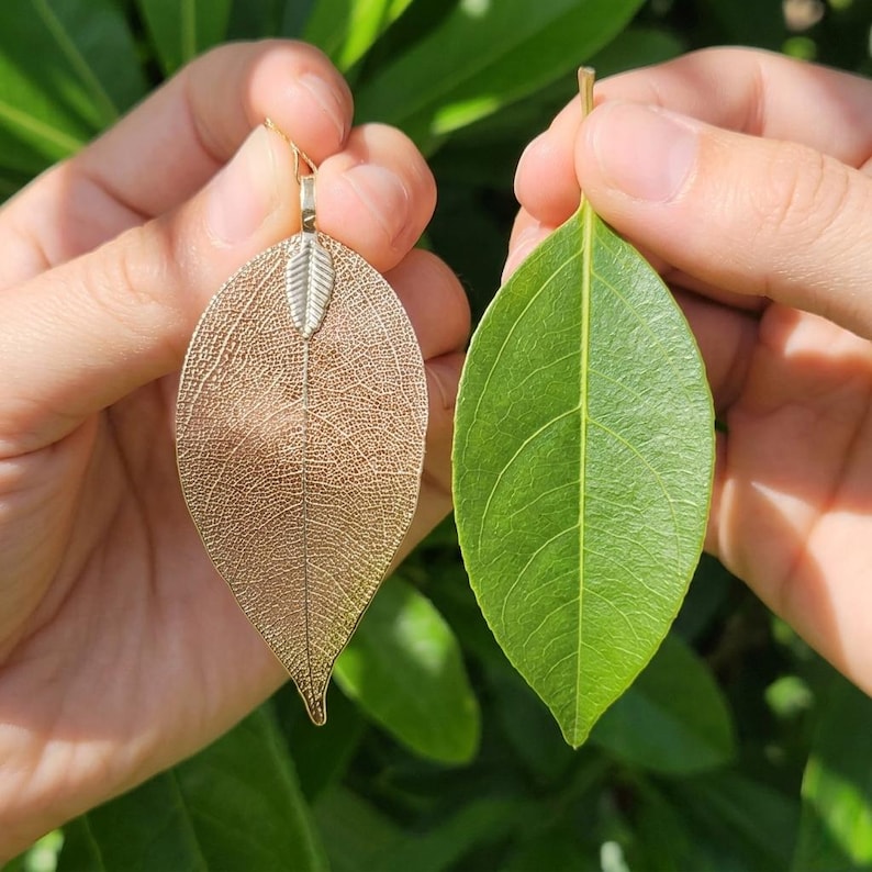 May include: A metallic gold leaf pendant featuring a detailed skeletal vein structure and a small silver-toned bail at the top, held alongside a natural green leaf of a similar shape and size to demonstrate the organic form and texture of the piece.