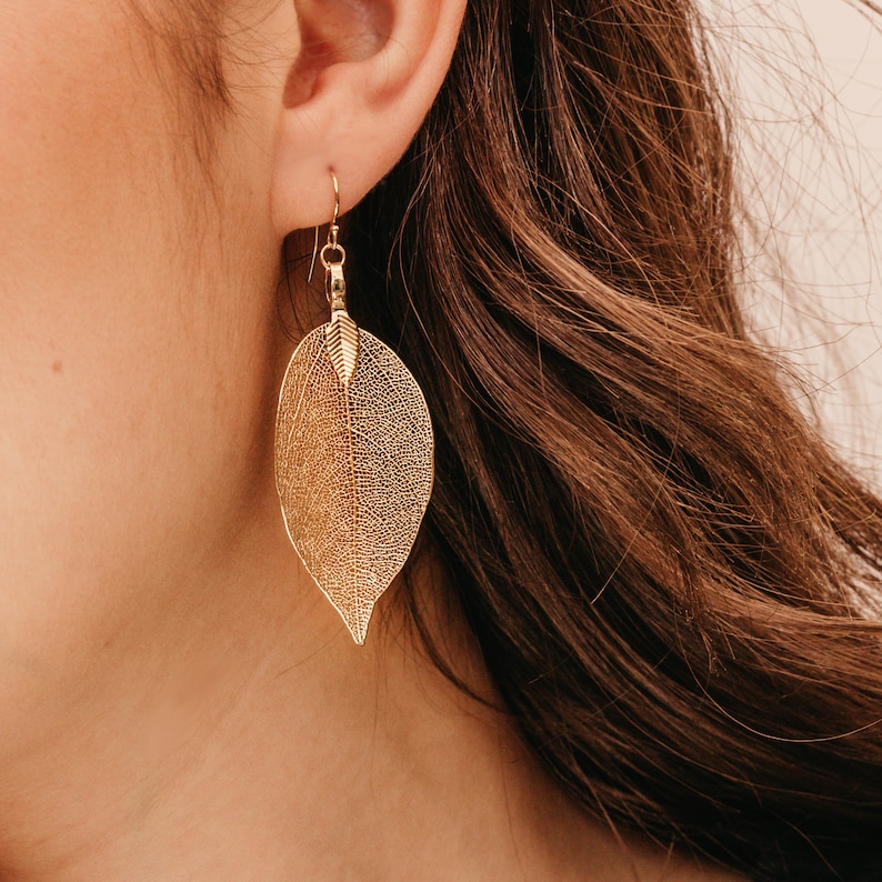 May include: A gold-toned drop earring shaped like a skeletonised leaf. The earring features a delicate, intricate network of veins forming the leaf structure, suspended from a simple gold hook wire that passes through the earlobe.