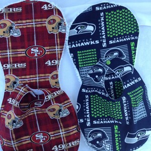 May include: Two baby bibs, one red and black with the San Francisco 49ers logo and the other blue and green with the Seattle Seahawks logo.