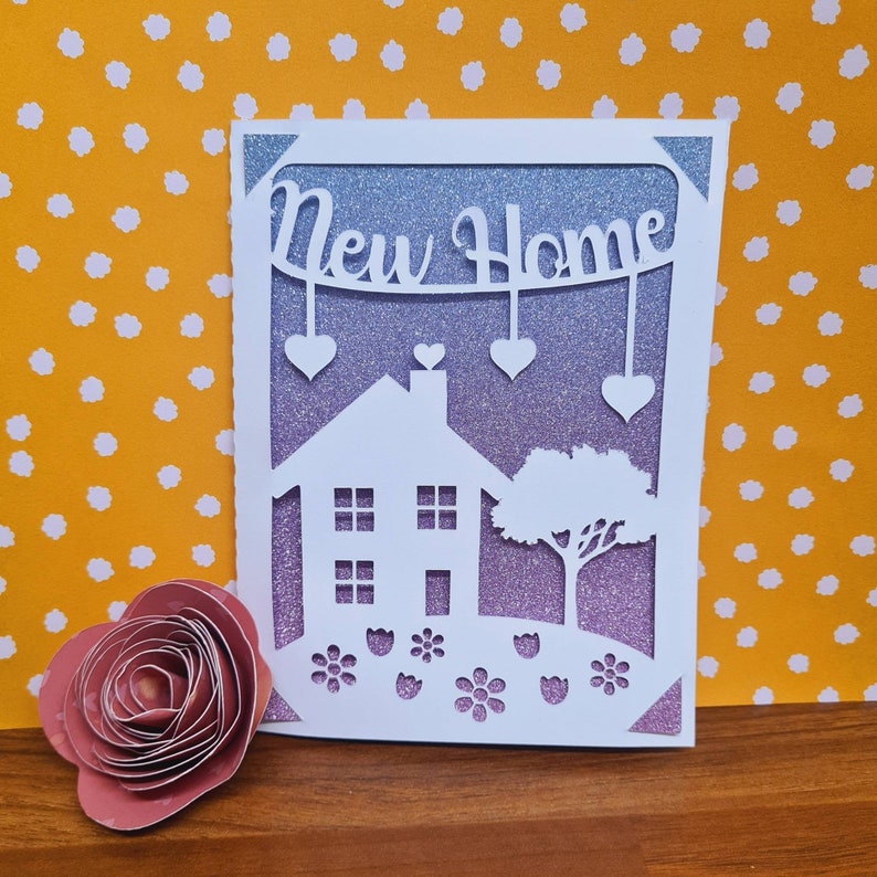 Pode incluir: Um cart&atilde;o branco com um fundo de purpurina roxo e um design recortado de uma casa, uma &aacute;rvore e flores. O cart&atilde;o diz "New Home" em uma faixa acima da casa.