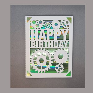 May include: White card with a cut-out design of gears and the words "HAPPY BIRTHDAY" on a holographic iridescent background.
