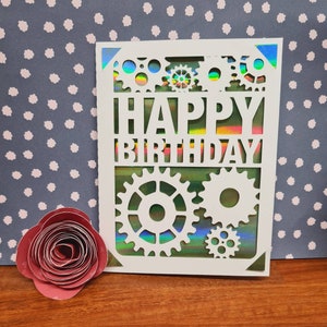 May include: White card with a holographic iridescent background and a cut-out design of gears and the words "Happy Birthday".