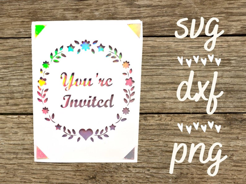 Puede incluir: Una tarjeta blanca con un acabado hologr&aacute;fico presenta una corona de flores y estrellas con el texto "You're Invited" en el centro. La tarjeta est&aacute; sobre un fondo de madera con las palabras "svg", "dxf" y "png" escritas en blanco.