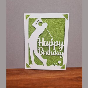 May include: A white card with a green glitter background. The card features a silhouette of a golfer swinging a club and the text "Happy Birthday".