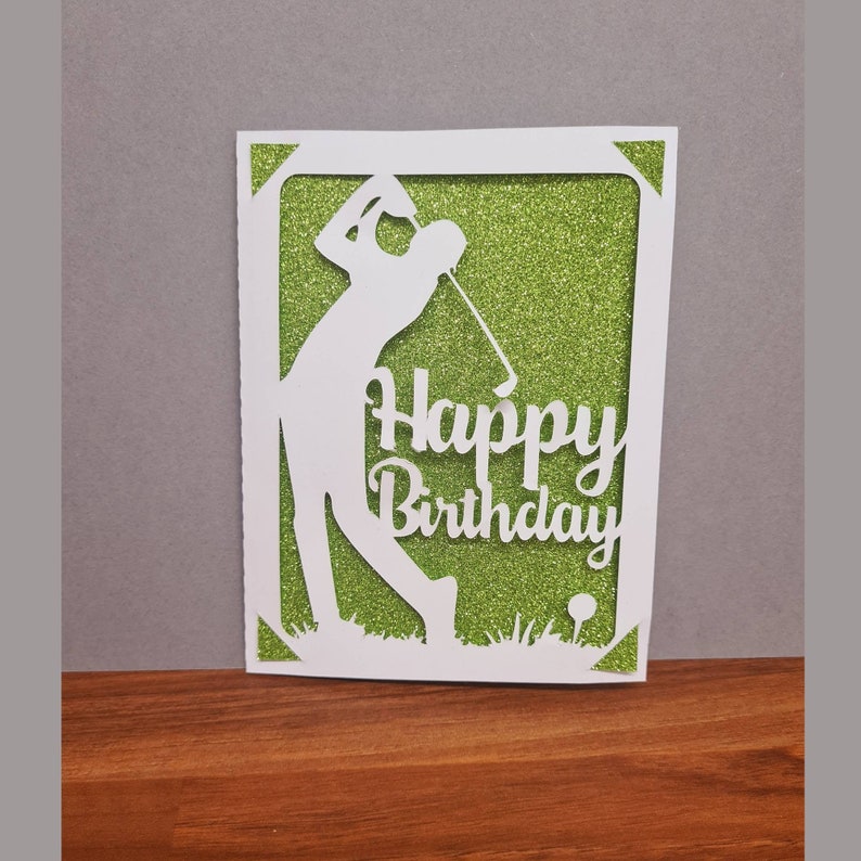 May include: A white card with a green glitter background. The card features a silhouette of a golfer swinging a club and the text "Happy Birthday".