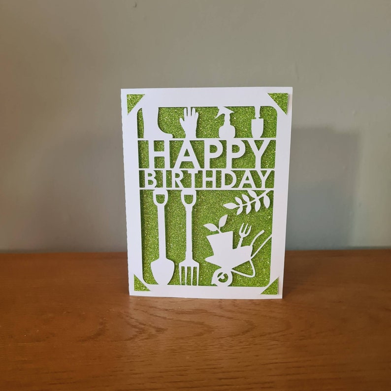 May include: White card with a green glitter background and a cut-out design of gardening tools and the words "Happy Birthday".