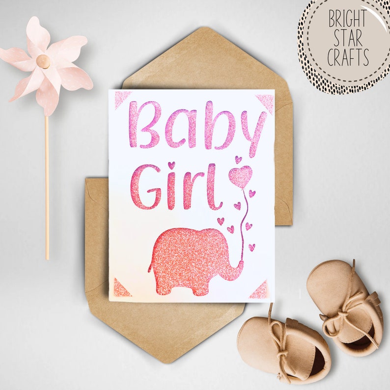 Puede incluir: Una tarjeta blanca con letras de purpurina rosa que dicen "Baby Girl". Debajo del texto hay un elefante de purpurina rosa con un globo rosa en la trompa. La tarjeta est&aacute; en un sobre marr&oacute;n.