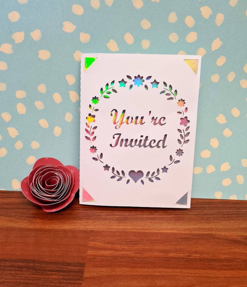 Puede incluir: Una tarjeta blanca con un dise&ntilde;o recortado de una corona floral con el texto "You're Invited" en el centro. La tarjeta tiene esquinas iridiscentes.