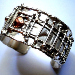 May include: A silver cuff bracelet with a copper heart design. The bracelet has a textured surface and a geometric pattern.