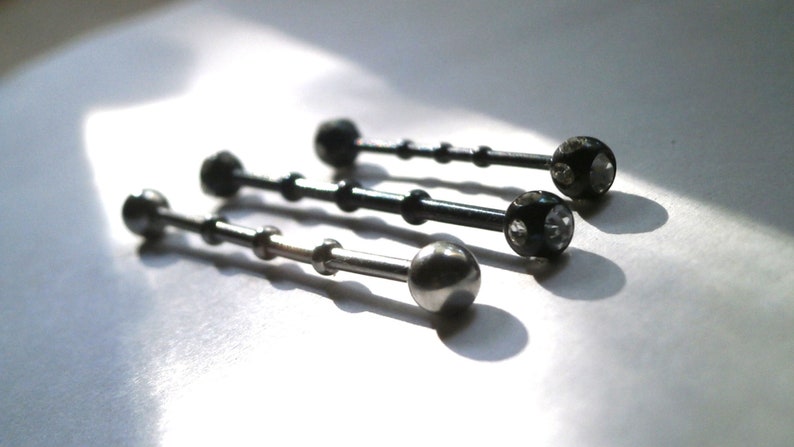 May include: Three silver barbell style body jewellery pieces with clear gem accents. The top two pieces are black with clear gems. The bottom piece is silver with a smooth ball end.